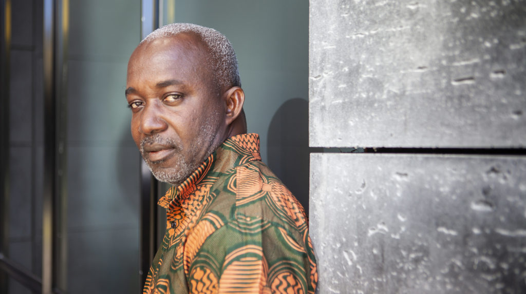 James Otto, kort grijs haar, oranje bloes, kijkt de camera in terwijl hij tegen de muur leunt.