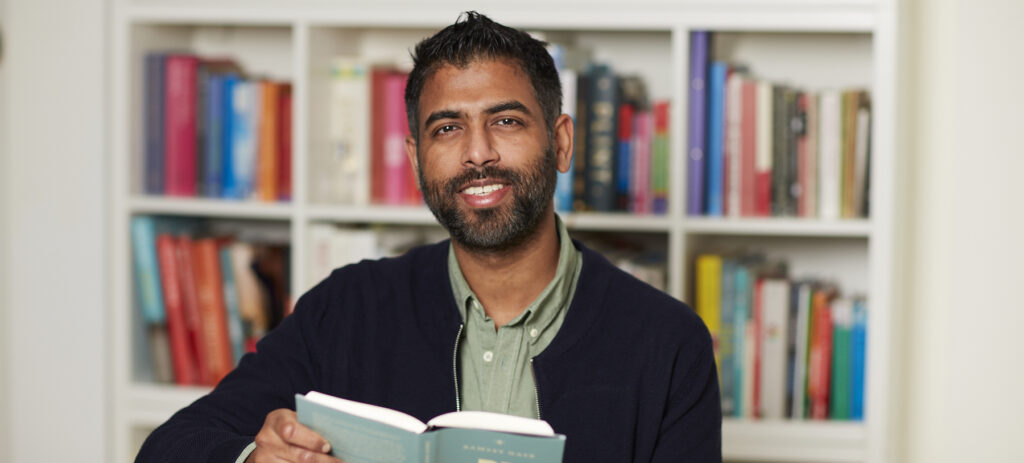 Een portret van Rakesh Kalloe. Hij zit voor een boekenkast, een boek open in zijn handen, en kijkt lachend de camera in. Hij heefty kort zwart haar en een stoppelbaard, en draagt een groen overhemd met een zwart vest.