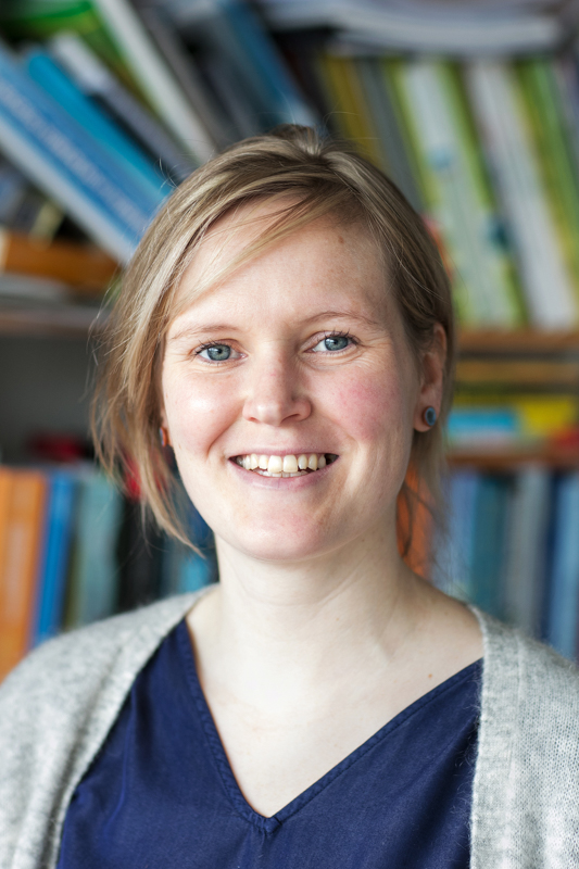 Portret van Ingrid Boas. Ze kijkt recht de camera in en lacht. Ze heeft rossig haar in een staart, een blauwe blouse en een grijs vest.