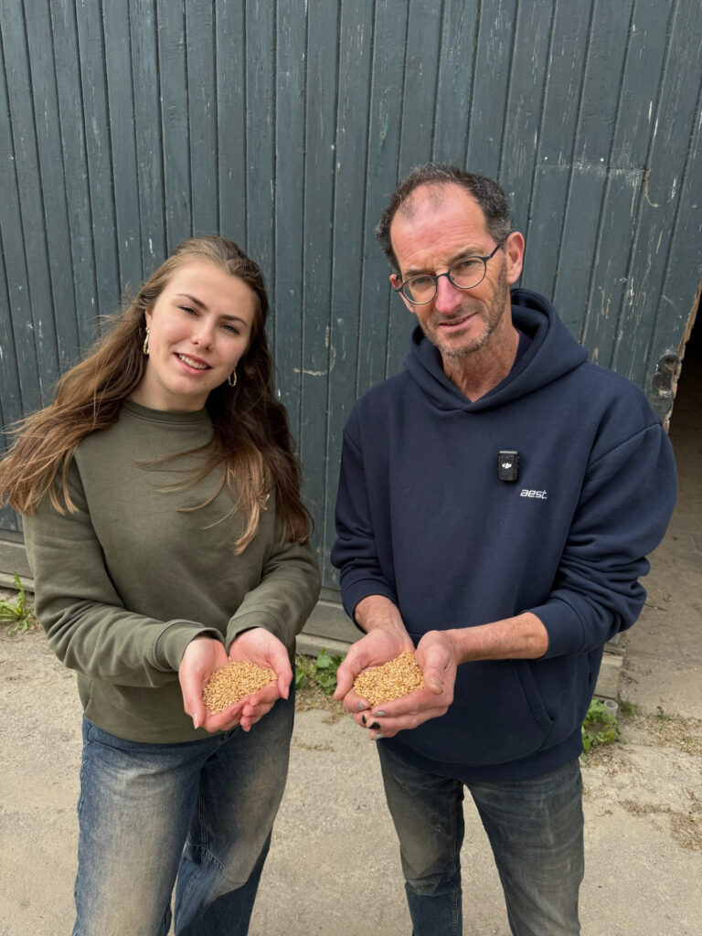 Twee mederwerkers, een jonge vrouw met groene trui en een oudere man met bril, laten de camera een handvol zaden zien.