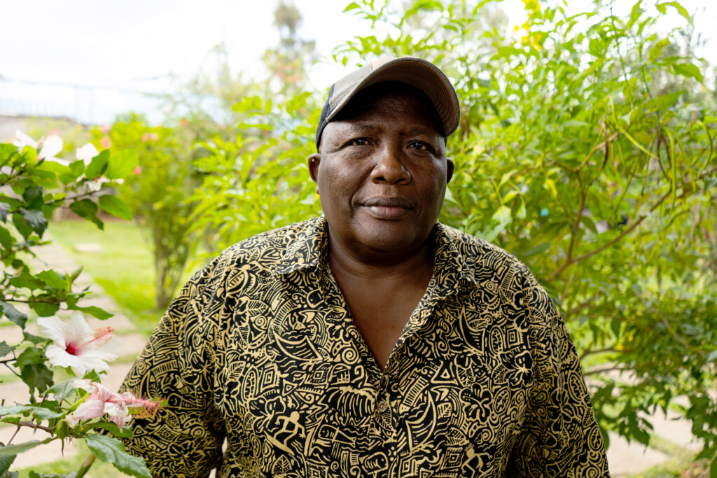 Portret van Meitamei Olol Dapash. Hij draagt een petje en een zwart wit gerpinte blouse, en kijkt recht de camera in.