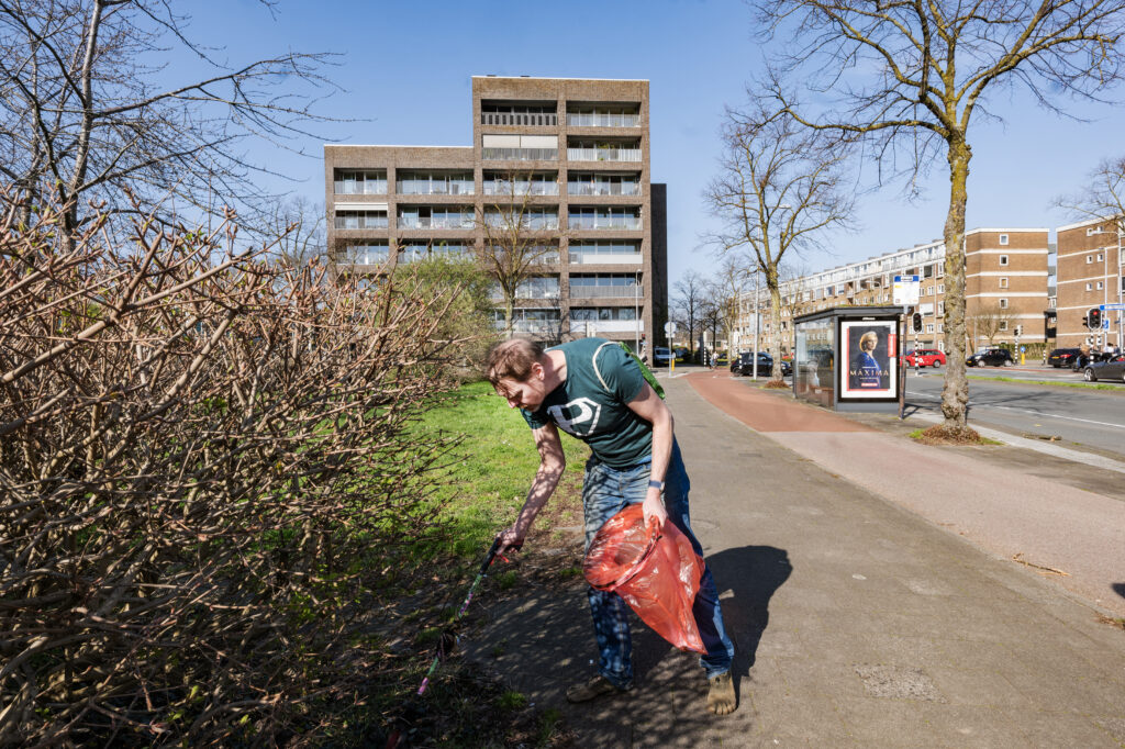 Anton zoekt onder een bosje naar zwerfafval. Hij heeft zijn rode vuilniszak en knijper gereed. Op de achtergrond staat een hoge flat.