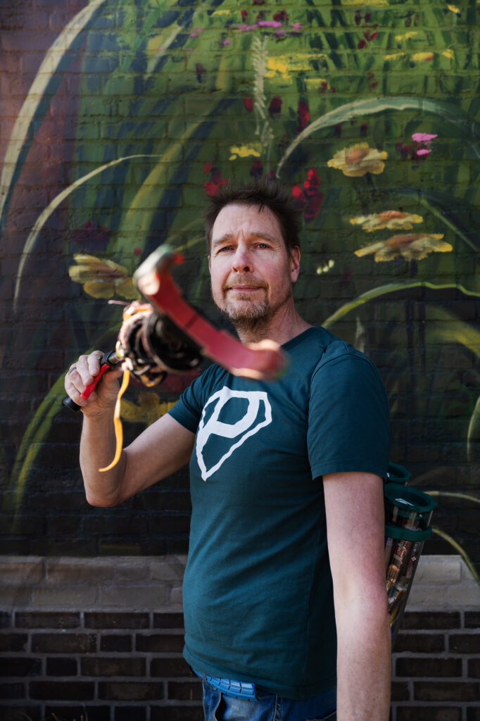 Anton kijkt vrolijk de camera in terwijl hij doet alsof hij met zijn knijper de camera grijpt. Hij heeft een groen 'plandelman' shirt aan (superman logo maar dan met de P). Achter hem op de muur staat graffiti van een bloemenveld