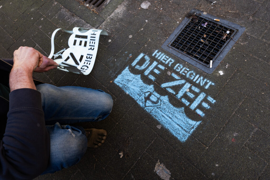 Anton spuit de zin 'hier begint de zee' in blauwe krijtspray naast de put. Onder de tekst staan blauwe golfjes met het plandelman logo.