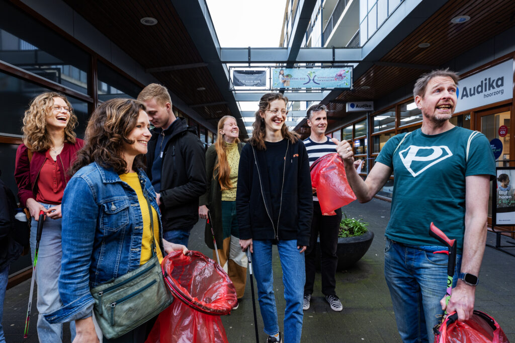 We zien een groep mensen die klaar staan om afval te prikken. Ze hebben allemaal en grote rode vuilniszak en een afvalknijper. We zien veel lachende gezichten terwijl Anton uitleg geeft. 
