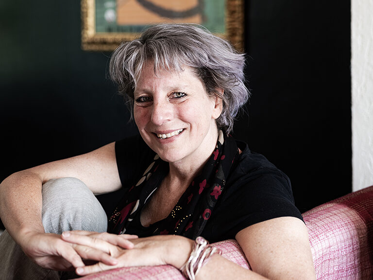 Frederike kijkt lachend de camera in vanaf de bank. Ze heeft half lang grijs haar, grijze ogen en draagt een zwart t-shirt en een zwarte sjaal met rode bloemen