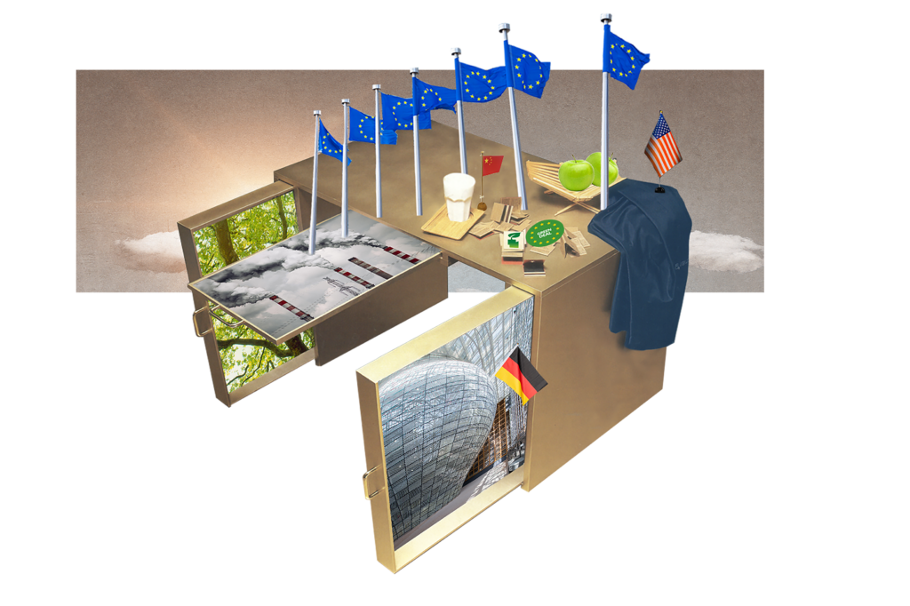 Een cartoon van een bureau met heel veel EU vlaggetjes, een vlag van de VS en een van china. Verder liggen er een volle koffiebeker, een appel, een jasje en een aantal papieren op het bureau. Op de zijkanten zijn plaatjes van bossen en fabrieksuitstoot geplakt.