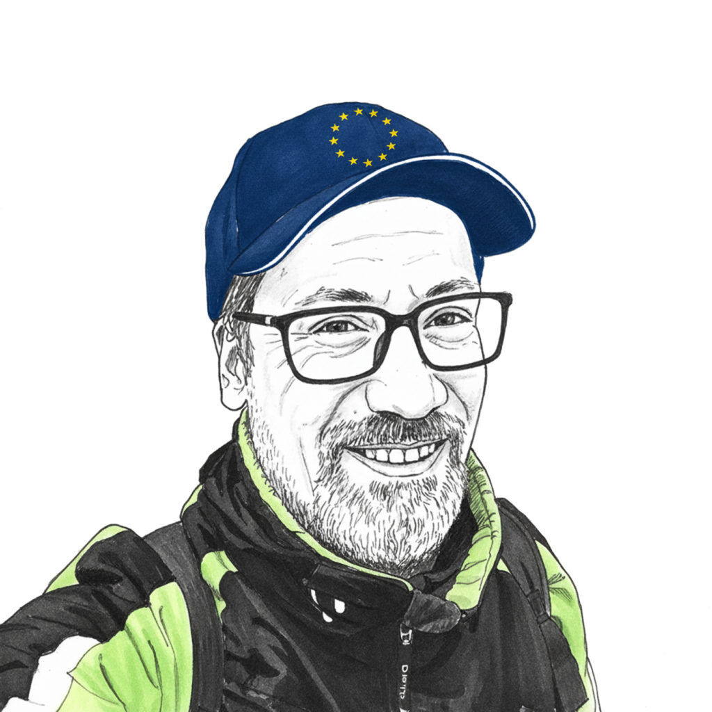 Een tekening van Hans Wetzels. Hij lacht de 'camera' in, draagt een petje met EU logo, bril en groen-zwarte jas. Hans heeft lachrimpeltjes, kort haar en een snor en baard.