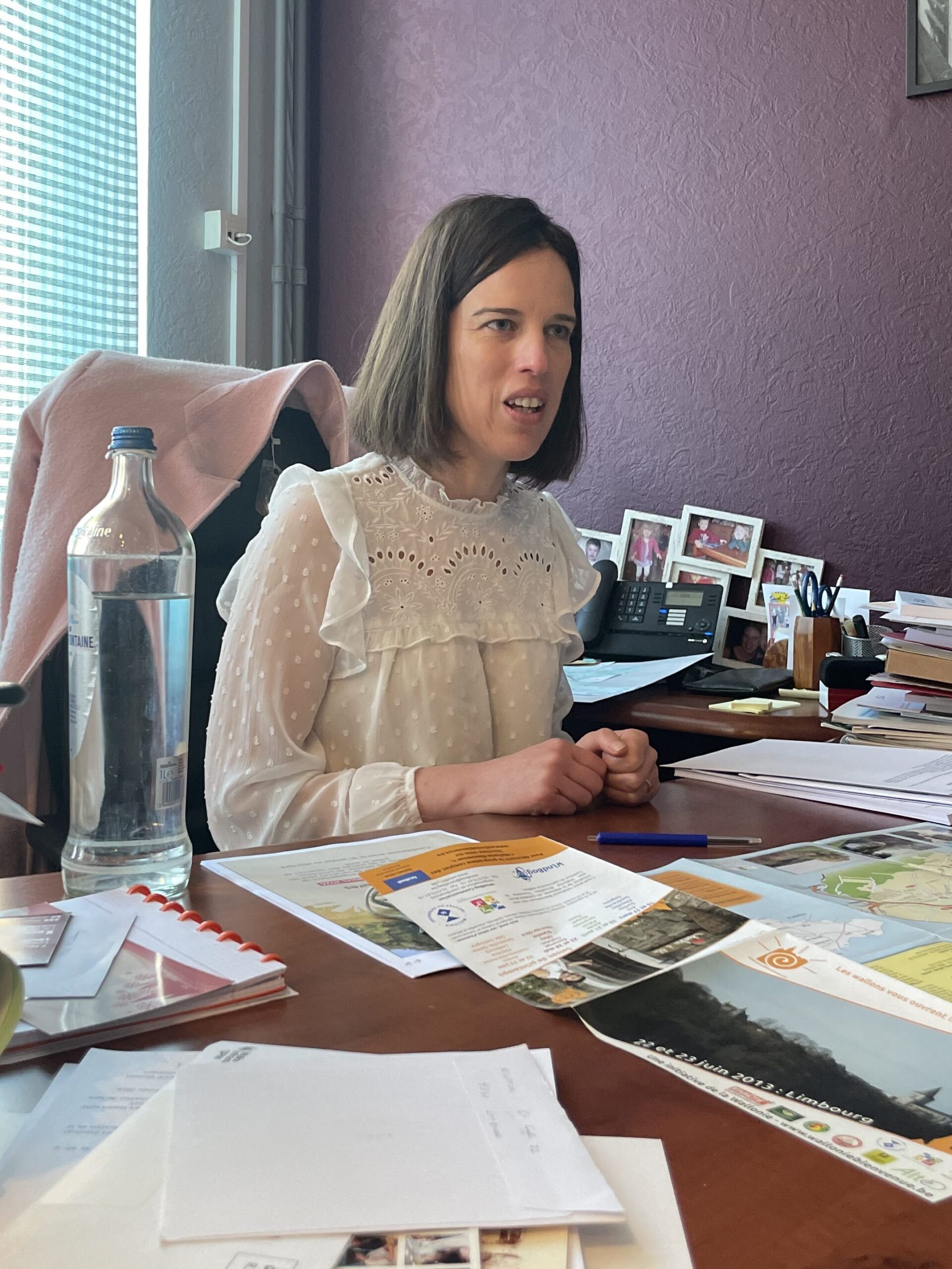 Valérie Dejardin zit achter een heel vol bureau. Ze kijkt serieus en lijkt midden in een gesprek. Ze heeft half lang, bruin haar een een witte bloes. Op haar bureau liggen veel papieren, een kaart, een aantal foto's en een waterfles. 