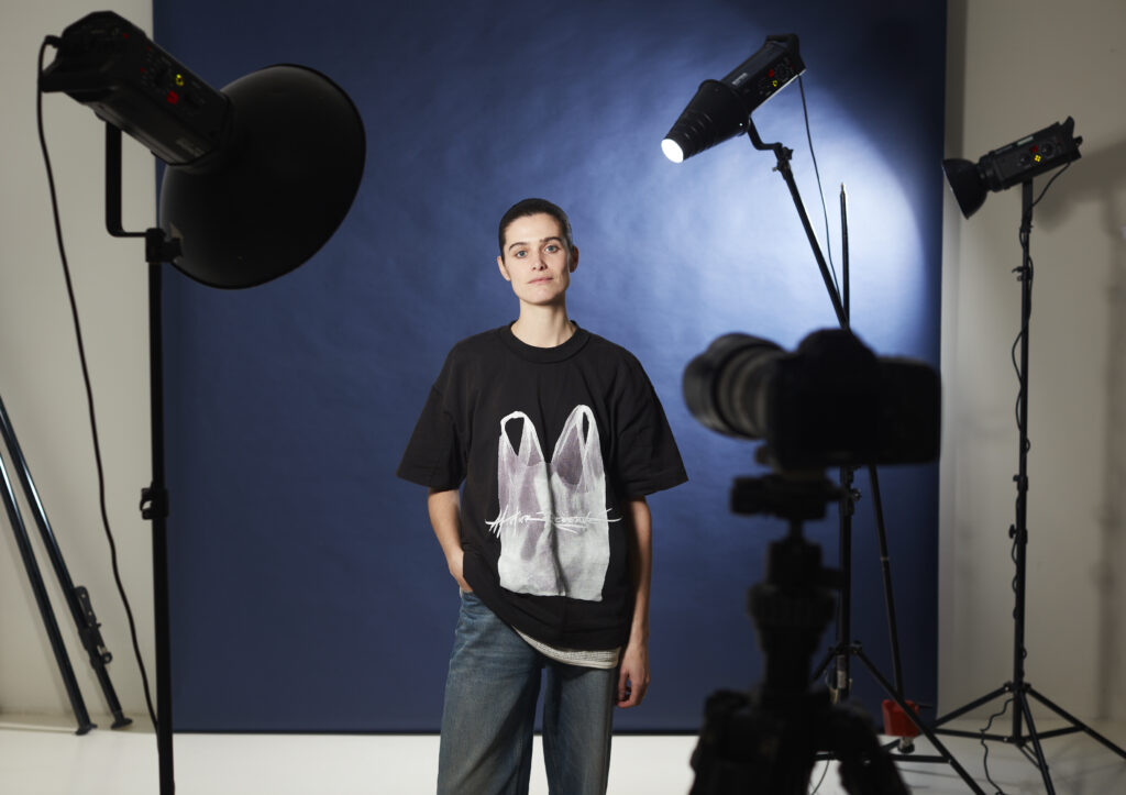 Kiki Boreel, kortgeschoren donker haar, t-shirt en jeans, kijkt recht de camera in. Ze staat voor een donker blauwe achtergrond. Om haar heen zien we camera's, verlichting en microfoons.