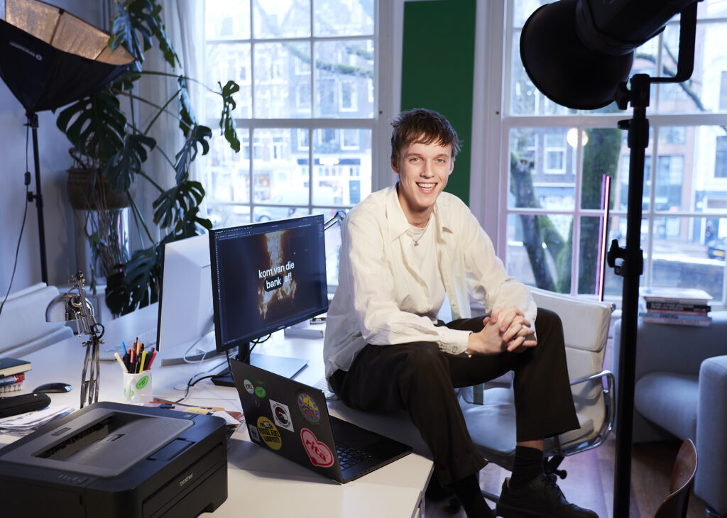 Morris vlijm zit op zijn bureau. We zijn in een lichte ruimte met grote ramen, de muur is groen en er staat een grote plant. Op het bureau staan computers en een printer. Morris draagt een wit overhemd over een wit t-shirt en een donkere broek en schoenen. Hij heeft kort blond haar en lacht de camera in. 