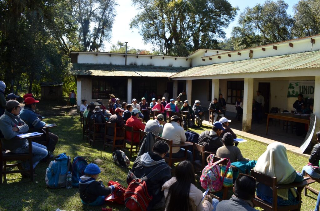 Een gemeenschapsvergadering in Tariquía. Mensen zitten in een halve cirkel op stoelen. Er zijn ook kinderen aanwezig. Achter ze staat een gebouw van 1 etage.