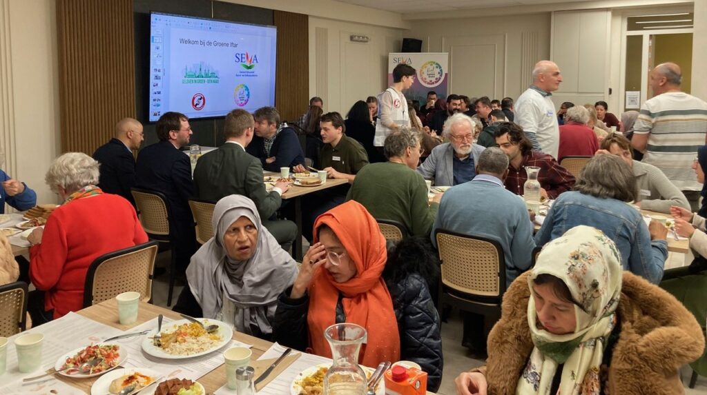 Een grote groep mensen zit aan de iftar in het Haagse Buurthuis. Ze zitten verdeeld over meerdere gedekte tafels. Of het digibord staan de logo's van de organisatoren.