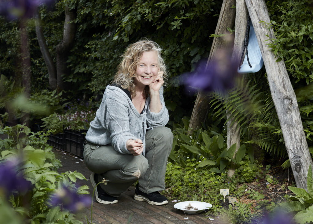 Anne Marie Faasen zit op haar hurken in de tuin en lacht de camera in. Naast haar liggen drie theezakjes op een bordje. Anne marie is een witte vrouw met blonde krullen, blauwe ogen, een grijs gebreid vest en grijze jeans. 