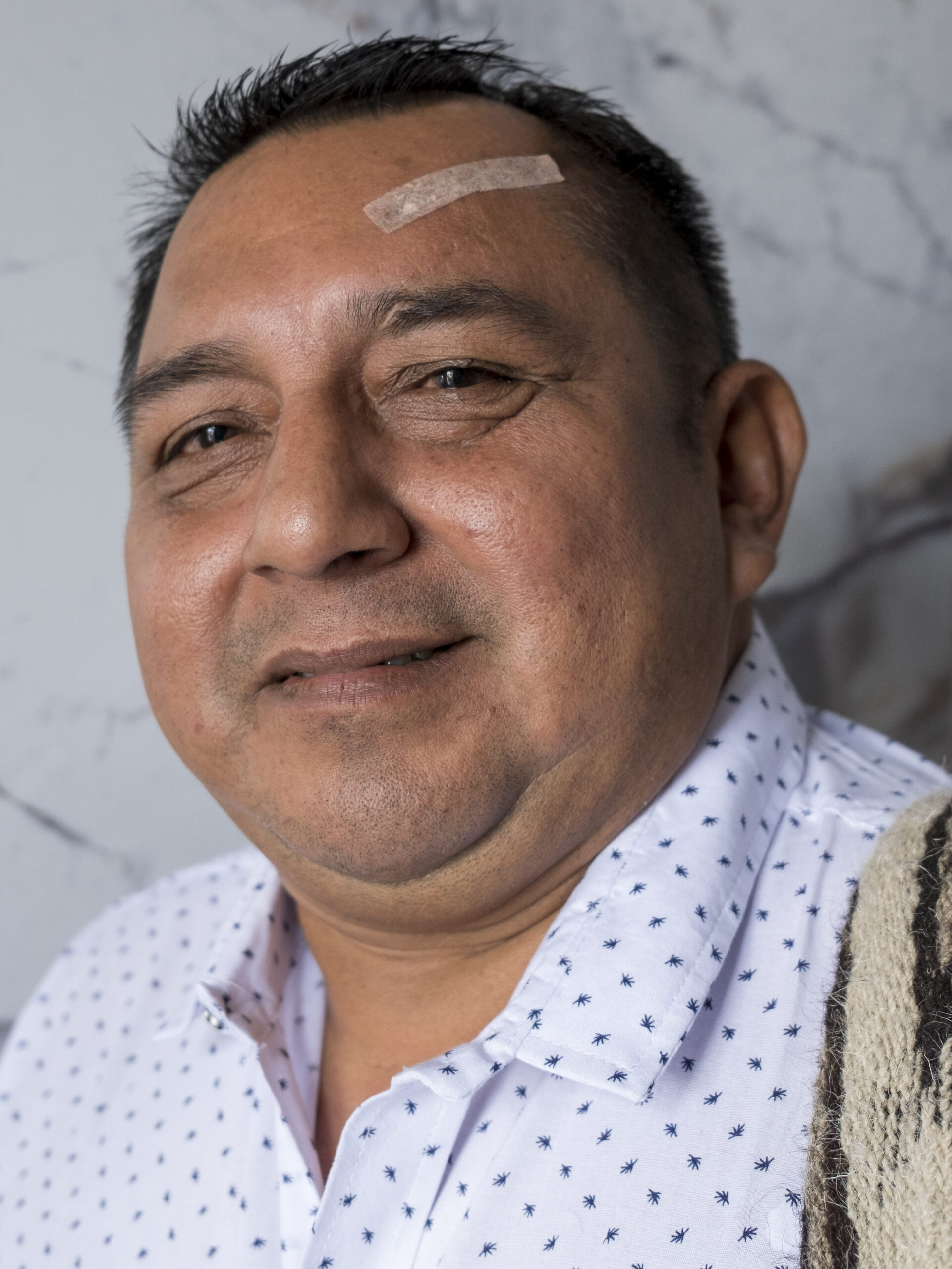 Antonio Matapí lacht de camera in. Hij Heeft kort zwart haar, een licht bruine huid en een pleister op zijn voorhoofd. Hij draagt een overhemd met donkerblauwe stippen en een tas over z'n schouder.