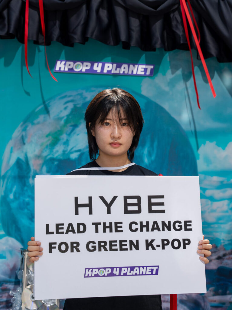 Dayeon bij een van de Kpop4Planet acties: ze houdt een bord vast met daarop: 'lead the change for green k-pop' en kijkt recht de camera in. Dayeon heeft half lang zwart haar met een pony en draagt een zwart t-shirt.