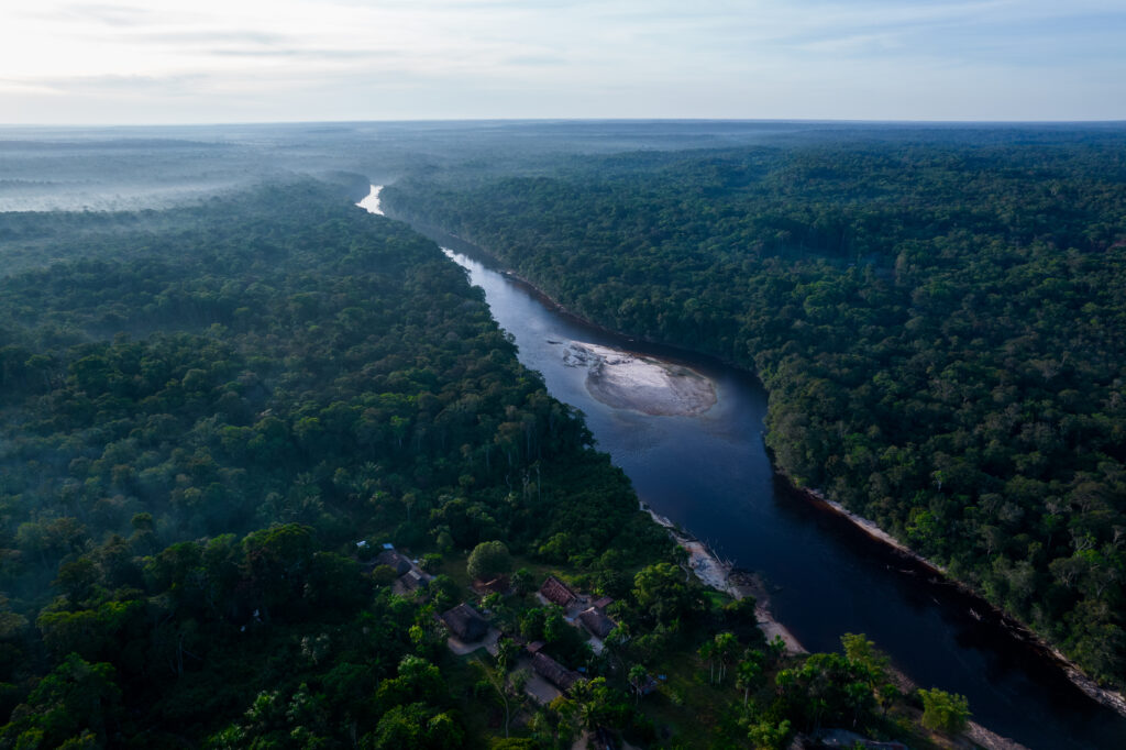 Een rivier stroomt door de prachtige Amazone. We kijken vanuit vogelvlucht over de ETI Pirá Paraná. Onder ons staan enkele huizen aan de oever van de rivier. 
