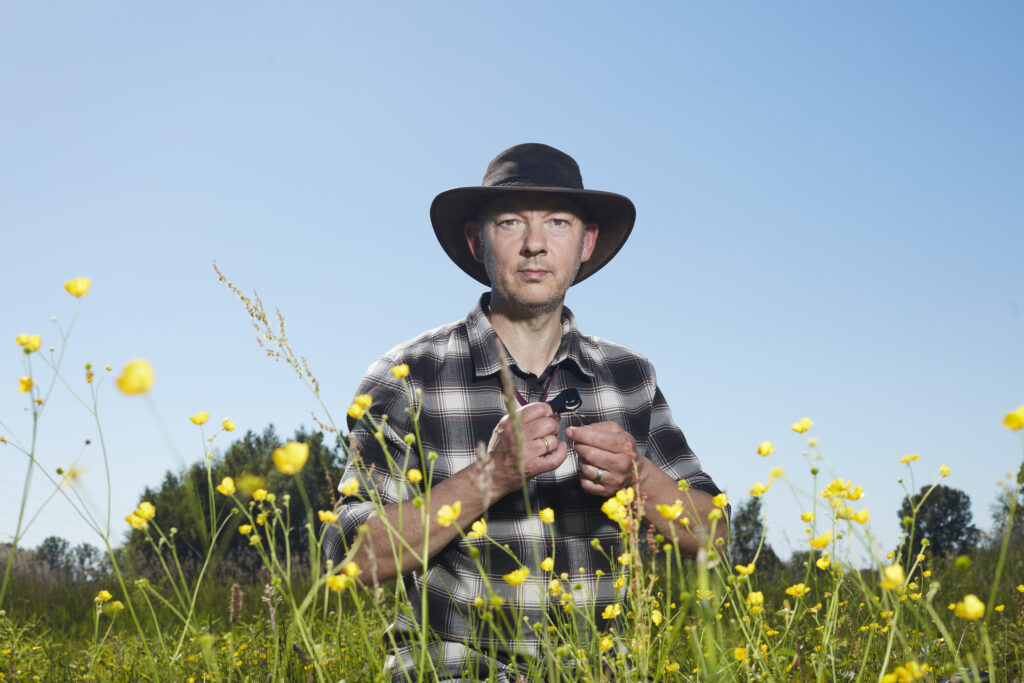 Gerrit staat midden in een weiland en kijkt recht de camera in. Hij draagt een grote hoed en een grijs-wit geblokt overhemd. Gerrit is een witte man met stoppels en een serieuze blik. Om hem heen bloeien de boterbloemen. 