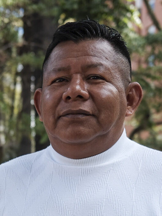 Gonzalo Macuna kijkt de camera in. Hij is een bruine man met kort zwart haar, bruine ogen en een wit t-shirt. Op de achtergrond zien we het bos. 