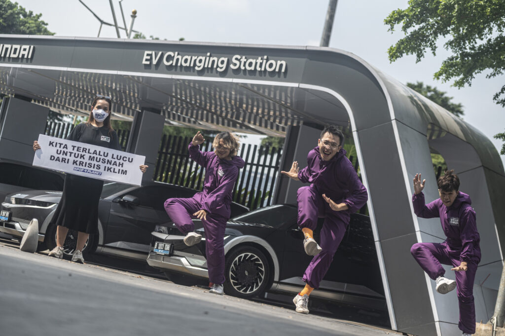 Een van de Kpop4Planet acties tegen Hyundai. Een aantal activisten danst in knalpaarse overalls voor een Hyundai laadstation. Een laatste houdt een protestbord vast, waarop in het Indonesisch staat: 'Wij zijn te schattig om dood te gaan door de klimaat crisis' 