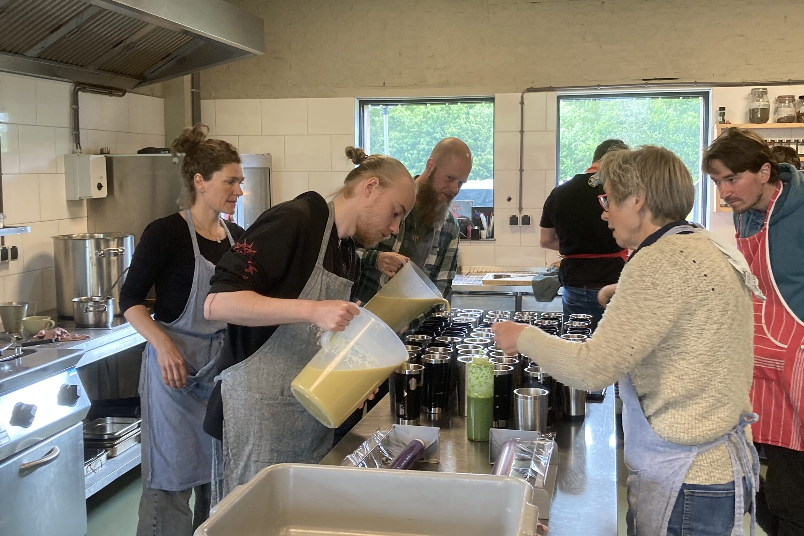 Een aantal mensen staat soep in thermosbekers te schenken. Ze hebben verschillende kleuren schorten aan en staan in een grote, professionele keuken.