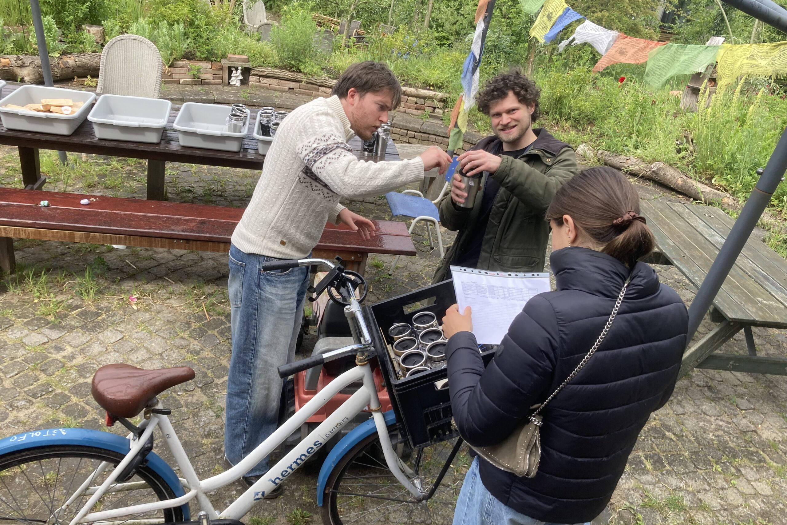 Drie mensen staan klaar oms oep door de buurt te fietsenw. Het fietskrat zit al vol met thermosbekers. Een persoon kijkt alvast op de lijst welke adressen die langs moet.