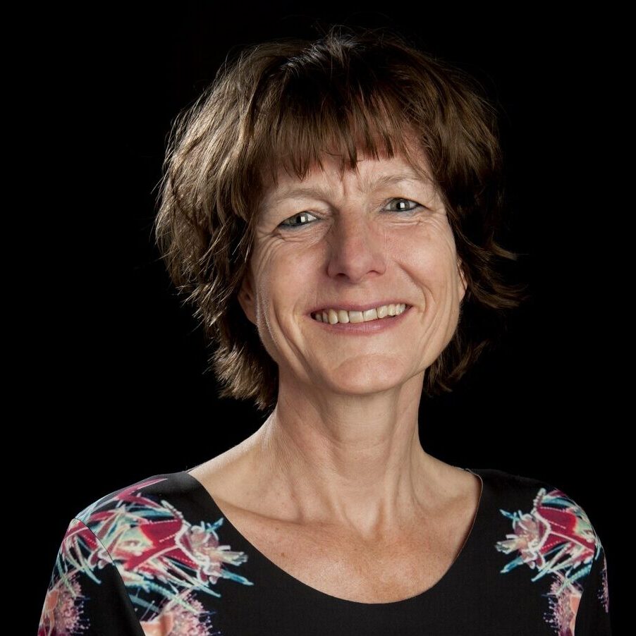 Linda Steg lacht de camera in. Ze is een witte vrouw met halflang bruin haar en bruine ogen. Ze draagt een zwart t-shirt met rood-blauwe bloemen erop.
