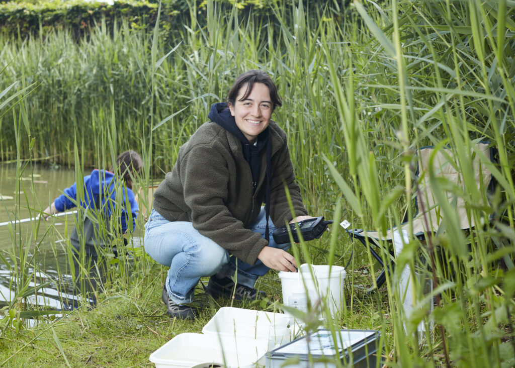 Lissy hurkt in het riet naast een sloot. Ze is een witte vrouw met donker haar en bruine ogen. Ze draagt bergschoenen, jeans een blauwe hoodie en een groen vest. Ze lacht recht de camera in en houdt meetapparatuur vast. Om haar heen staan emmers en een kampeer stoeltje. 