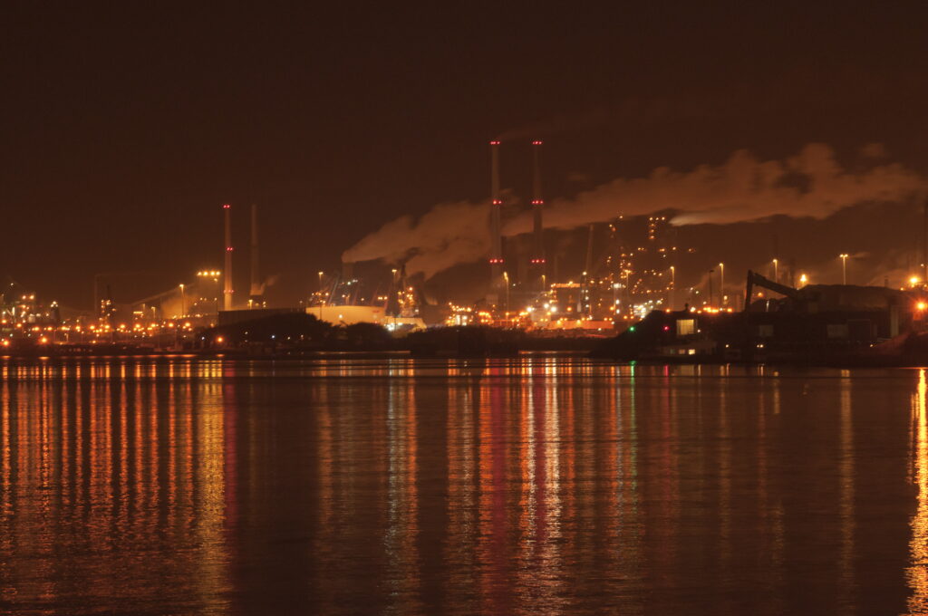 Een panorama van TATA steel s'nachts. Enorme rookwolken komen uit de schoorstenen en de hele lucht en het water gloeit oranje van de hoeveelheid lampen. 
