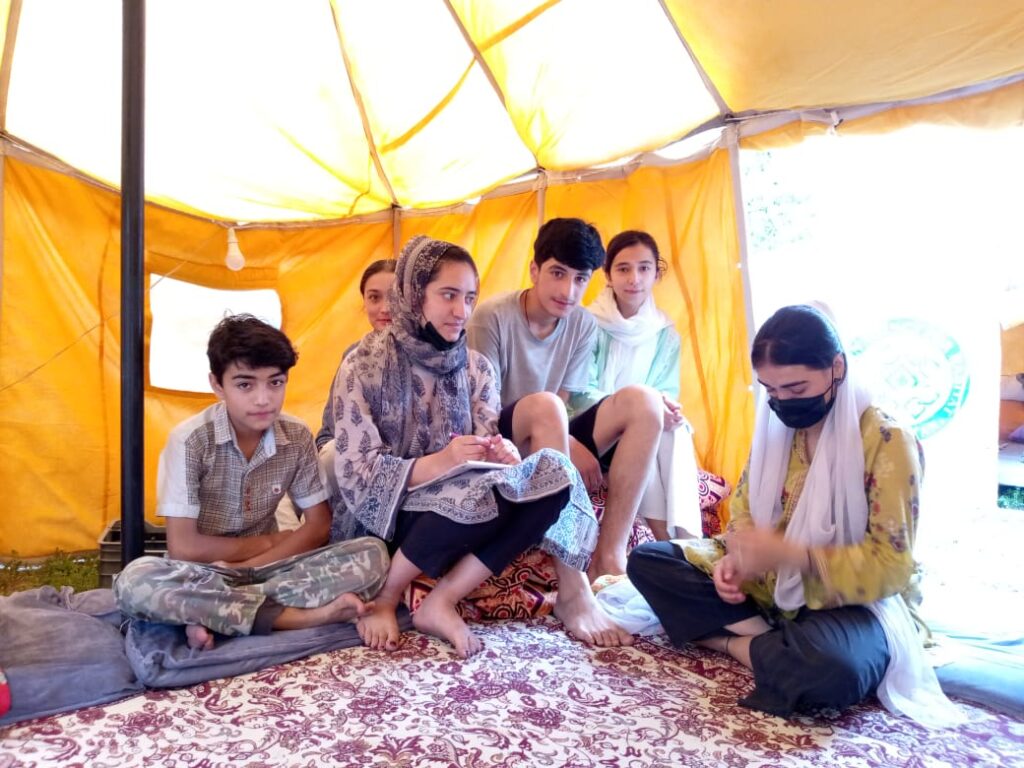 De familie van Zahid Mehmood in de tent waar ze wonen sinds hun huis is verwoest. Ze zitten met zijn zessen op kussens midden in de knalgele tent. Er ligt een rood-wit tapijt op de vloer. Alle vier de vrouwen dragen hun haar naar achter gebonden en dragen sjaal om hun nek. Een vrouw daagt een blauw-witte hoofddoek met een gedetailleerd patroon. De jongens hebben kort zwart haar en kijken recht de camera in.