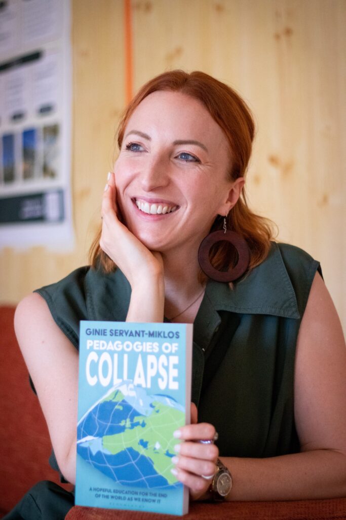 Ginie Servant-Miklos houdt haar nieuwe boek vast: Pedagogies of Collapse. Ze draagt een groene blouse en grote houten oorbellen. Haar hoofd steunt op haar hand en ze kijkt lachend langs de camera.