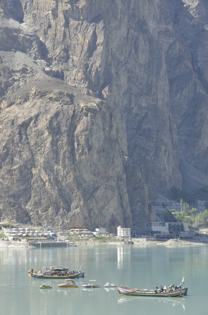 Toerisme bij het Attabd-meer in Gilgit-Baltistan. Op een meer varen meerdere felgekleurde bootjes. Achter het meer verrijst een enorme bergwand, met op de oever een aantal huizen.