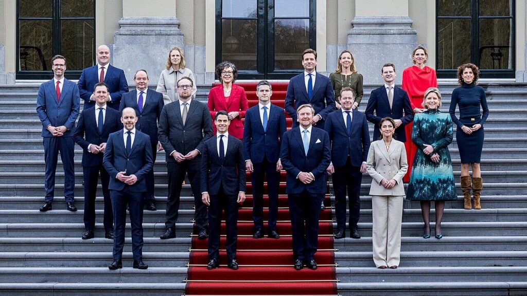 Bordesfoto van kabinet Jetten. Alle nieuwe ministers staan rondom de koning op het bordes van het paleis. Iedereen draagt professionele kleding en kijkt recht de camera in met een glimlach. 