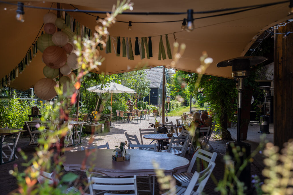 Sfeervol plaatje van het terras op de Eemlandhoeve. Alles staat onder een luifel, met ronde tafels en stoelen eromheen. Boven het terras hangen lampjes.