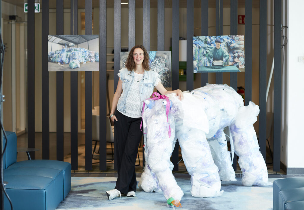 Evelyn Brakema staat in een kantoorruimte bij een grote olifant, gemaakt van plastic afval, dat is vrijgekomen bij medische behandelingen. Op de achtergrond hangen 3 foto's van medisch afval. Evelien is een lange witte vrouw, met lang krullend bruin haar. Ze draagt een wit tshirt met spijkerjasje en zwarte broek, en kijkt lachend de camera in terwijl ze op de olifant leunt. 