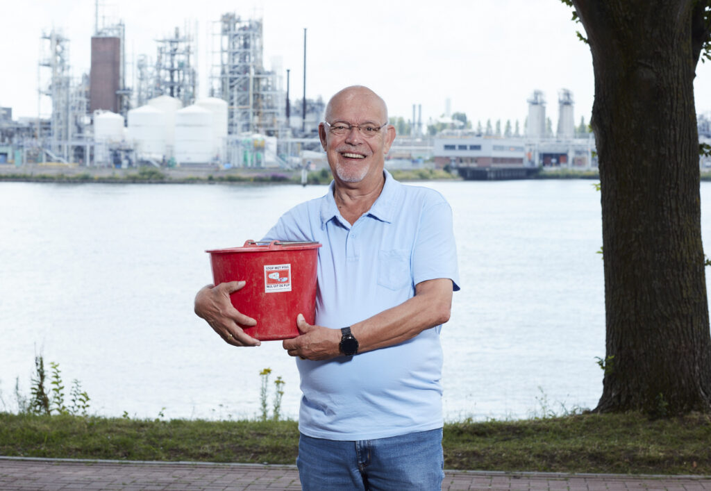 Kees van der Hel staat buiten, met een rivier en de Chemoursfabriek op de achtergrond. Hij houdt een rode emmer vast. Kees is een kale witte man met een bril. Hij draagt een lichtblauwe polo en een blauwe spijkerbroek.