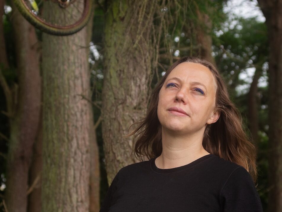 Lisa Doeland staat in een bos met dennenbomen. In een van de bomen hangt een oude, oranje fiets