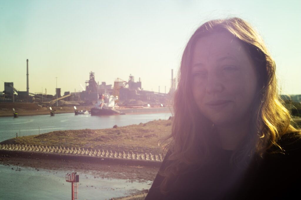 Lisa Doeland kijkt in de camera, met de zon in haar rug. Op de achtergrond is Tata Steel te zien.