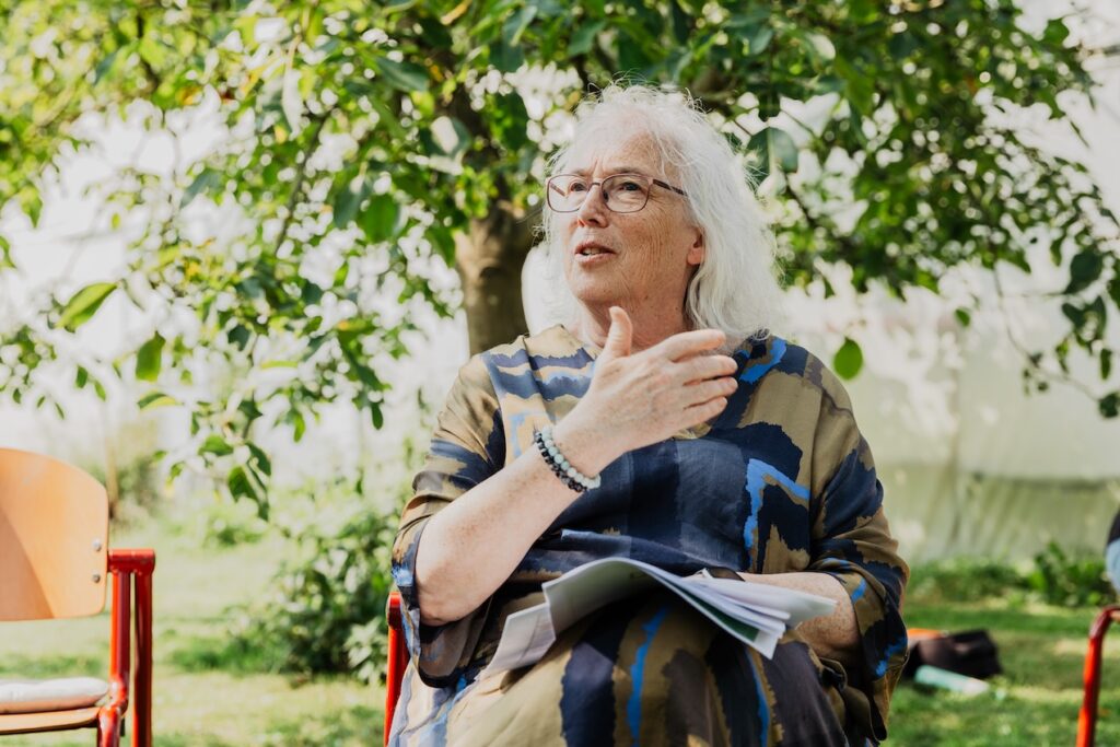 Sandra Ball zit buiten op een stoel voor een boom. Rechts en links van haar staan ook stoelen. Sandra is een witte vrouw met lang grijs haar, een bril en draagt een wijde blouse. 