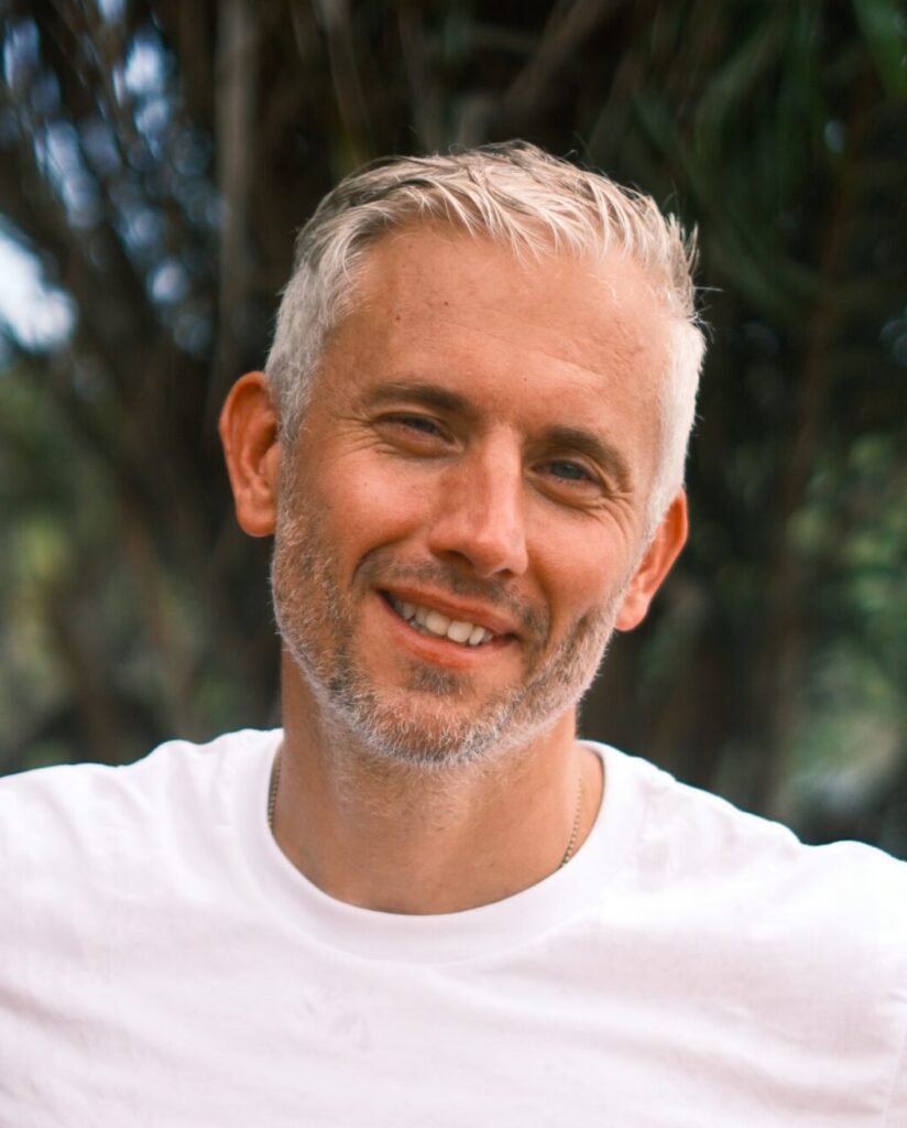 Portret van Tim van Deursen. Tim is een witte man met kort grijs haar wat opzij gekamd zit. Hij draagt een effen wit t-shirt en lacht de camera in. 