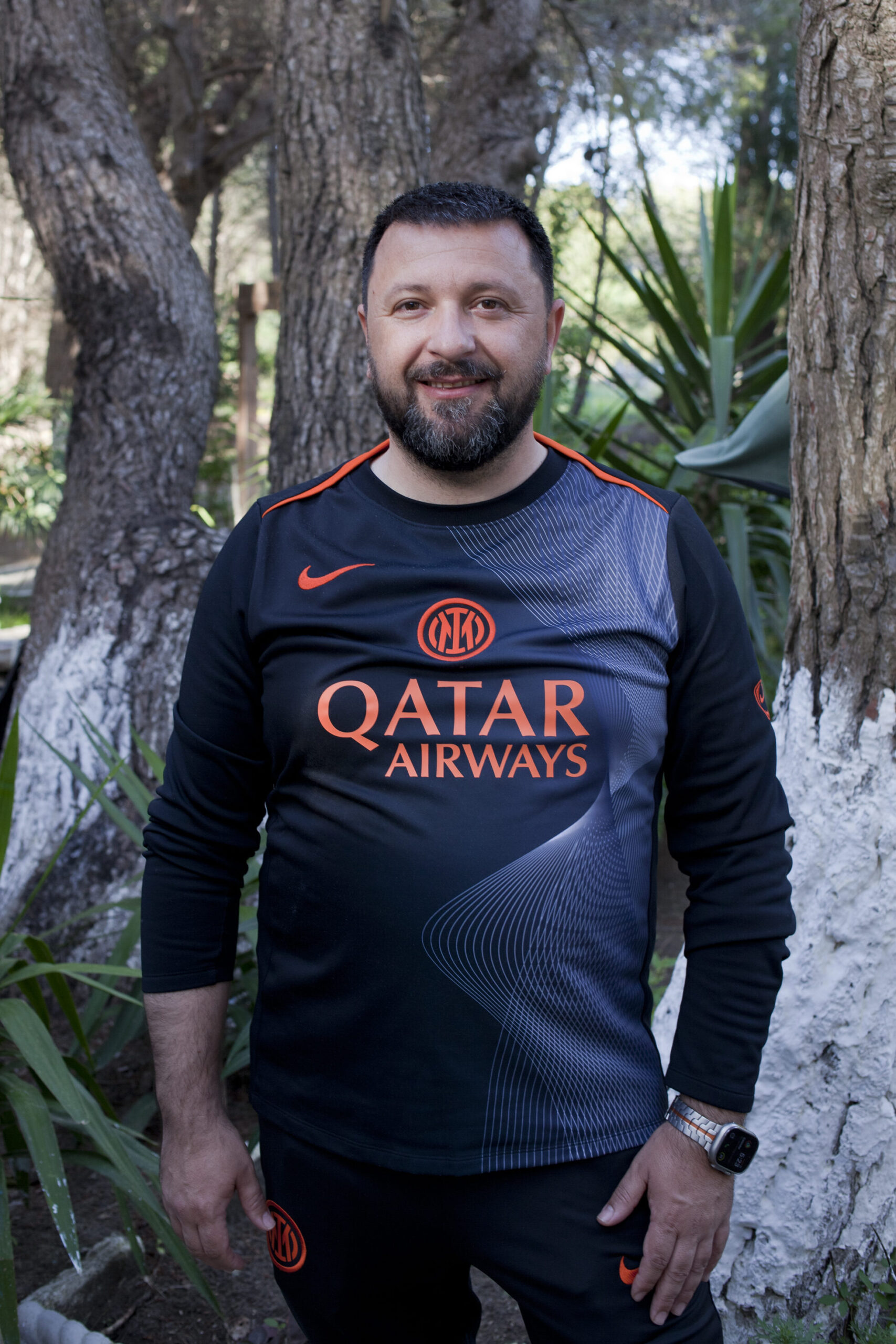 Olsi Nika lacht de camera in. Hij draagt een zwart-oranje voetbal shirt en een zwarte broek. Olsi heeft een lichte huid, kort zwart haar en een korte zwarte baard. 