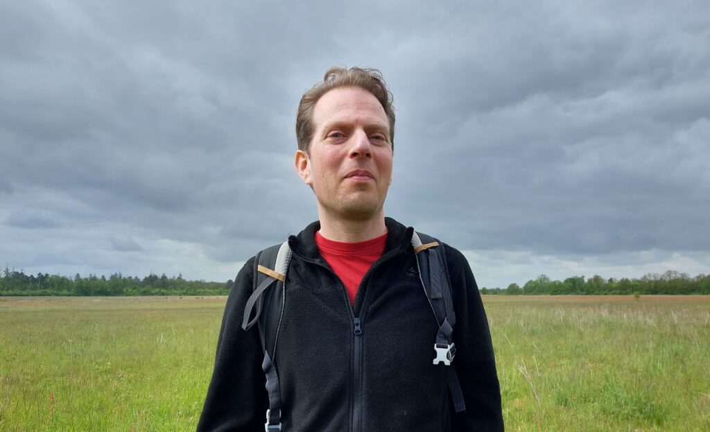 Een foto van Machiel van Veen, vanaf zijn middel. Hij staat in een groot uitgestrekt weiland met een dreigende wolkenlucht. Machiel is een witte man met kort bruin haar. Hij draagt een rood shirt, zwart vest en rugzak en kijkt recht de camera in.