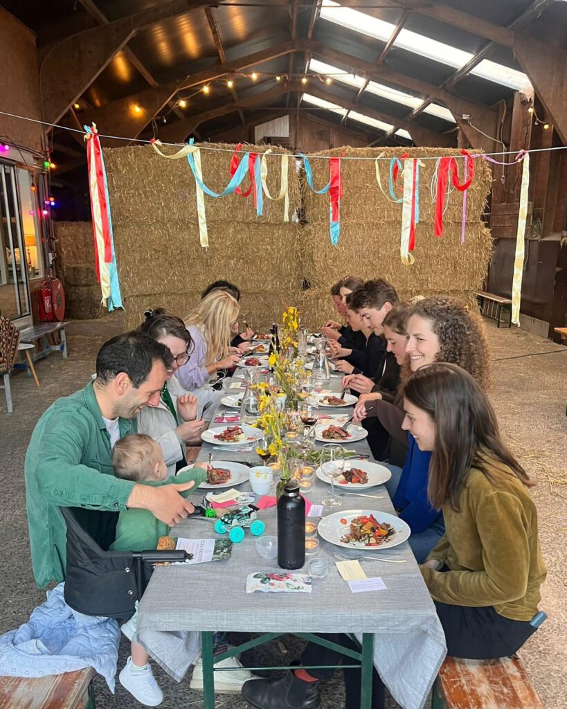 Een diner van Eigen Krachtvoer in een schuur. Een groep van rond de tien mensen zit aan een lange gedekte tafel samen te eten. Er staan gele bloemen op tafel, er hangt een kleurrijke slinger en op de achtergrond liggen hooibalen.