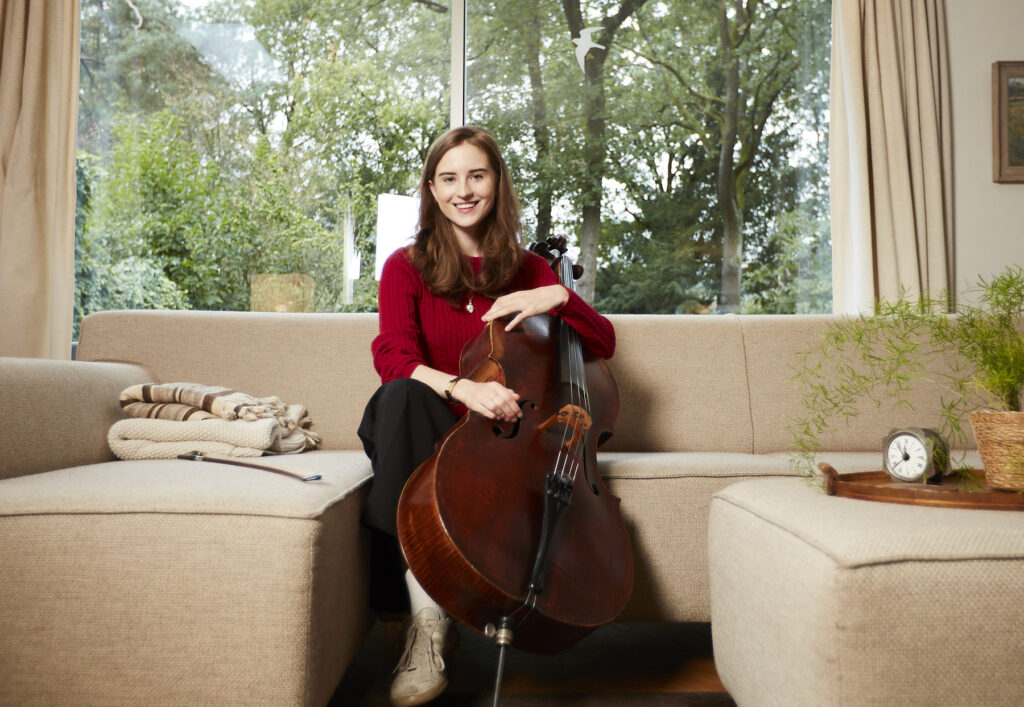 Donna zit met haar cello op de bank. We zien haar echt van voren, ze lacht de camera in. Donna is een witte meid met lang bruin haar. Ze draagt een knalrood lange mouwen shirt, een zwarte broek en witte schoenen. 