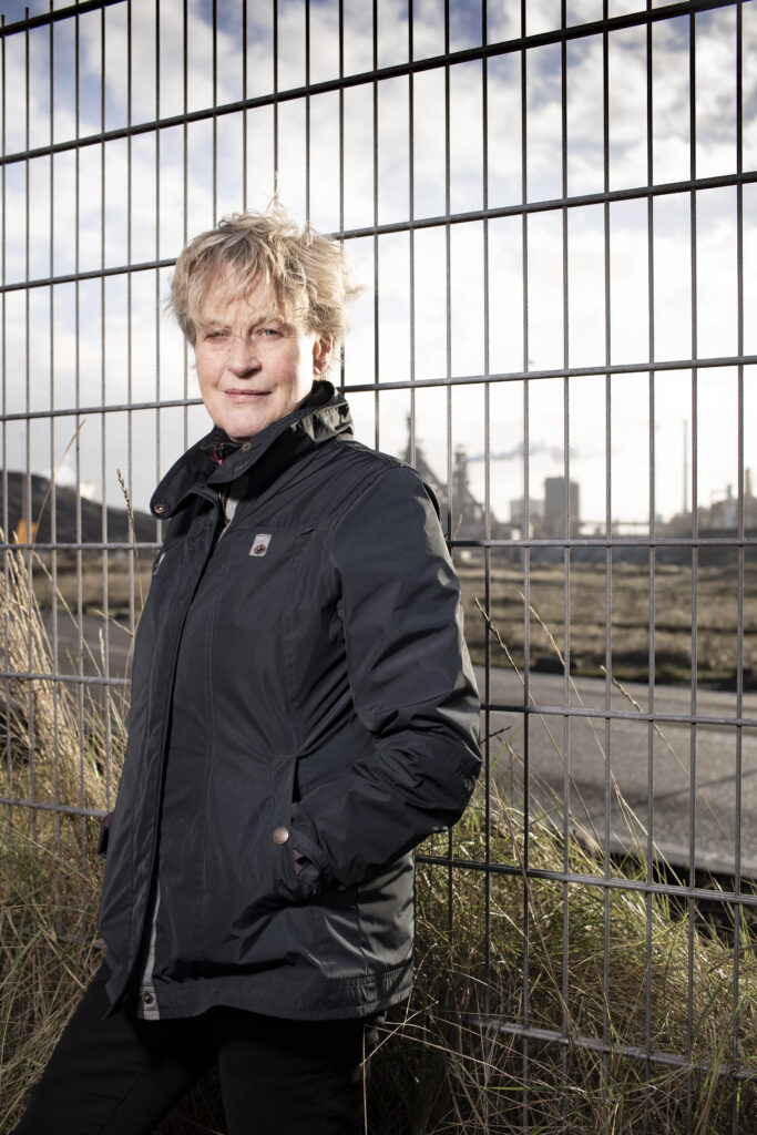Bénédicte Ficq leunt tegen het hek van tata Steel. Ze draagt dezelfde zwarte jeans en grijze jas, en kijkt recht de camera in.