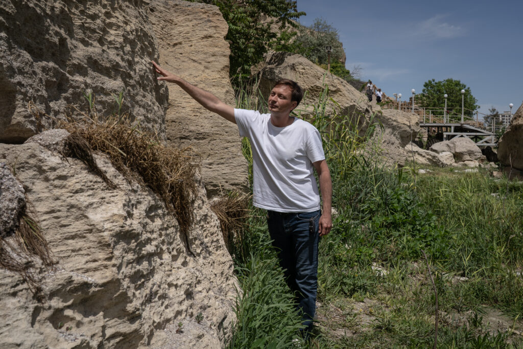 Kirill Ossin wijst op droogliggend gesteente aan tot waar het water kwam in 1999. Hij staat op de nieuw begroeide grond, en wijst een stukje boven zijn hoofd. 