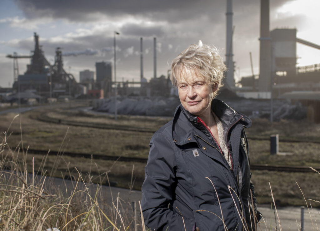 Bénédicte staat in hoog gras en kijkt stoer de camera in. Ze is een witte vrouw met kort blond haar en draagt zwarte jeans en een grijze jas. Op de achtergrond doemt TATA Steel op.