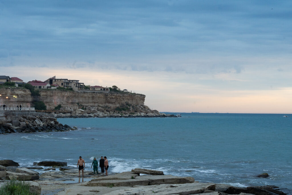 De kust van Aktau. Kliffen steken af tegen de lucht, het water is fel blauw. Mensen staan op het droogliggende strand, klaar om te zwemmen. 