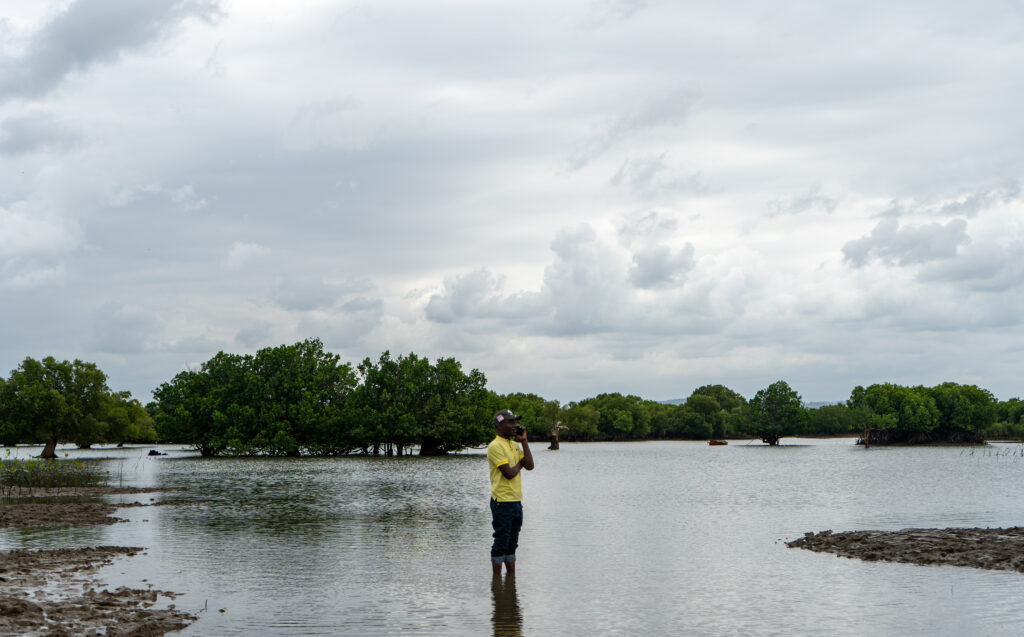 Mbaarak Abdalla staat in de baai tussen de mangrovebossen te bellen. Mbaarak is een zwarte man met kort zwart haar. Hij draagt een gele polo, groen petje en opgestroopte spijkerbroek. 