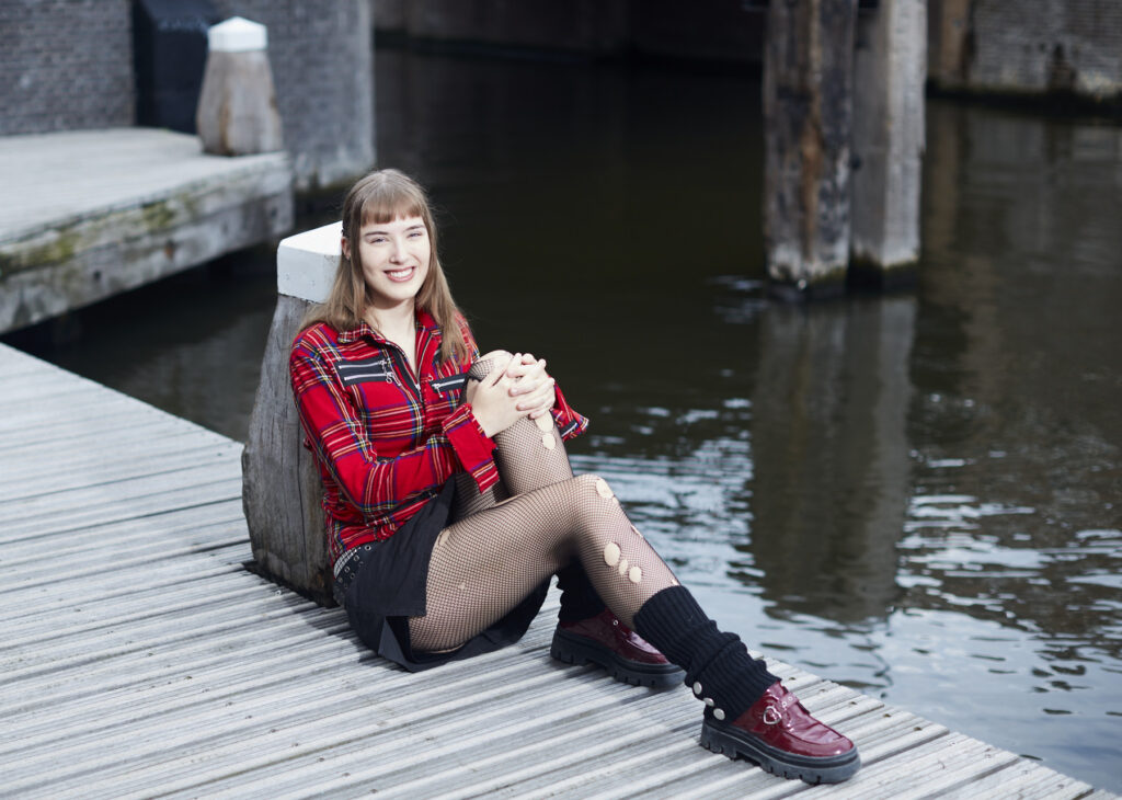 Merel zit op een pier en leunt tegen een paal aan. Merel is een witte meid met lang bruin haar en en pony. Ze draagt een rood geblokt overhemd, een zwarte rok, zwarte panty en rode schoenen